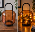 Amber glass decorative lantern with black metal handle, shown plain and styled with warm fairy lights